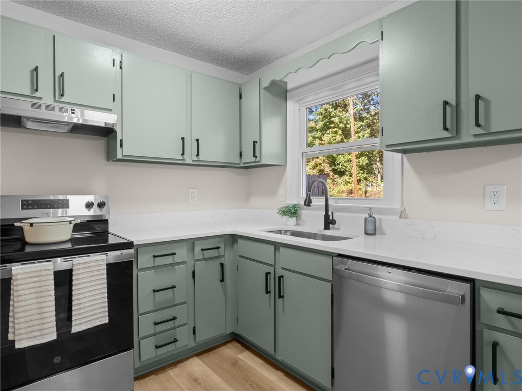 Kitchen featuring sage green shaker-style cabinetry, matte black hardware, white solid-surface countertops, stainless steel appliances, and wood-finish flooring