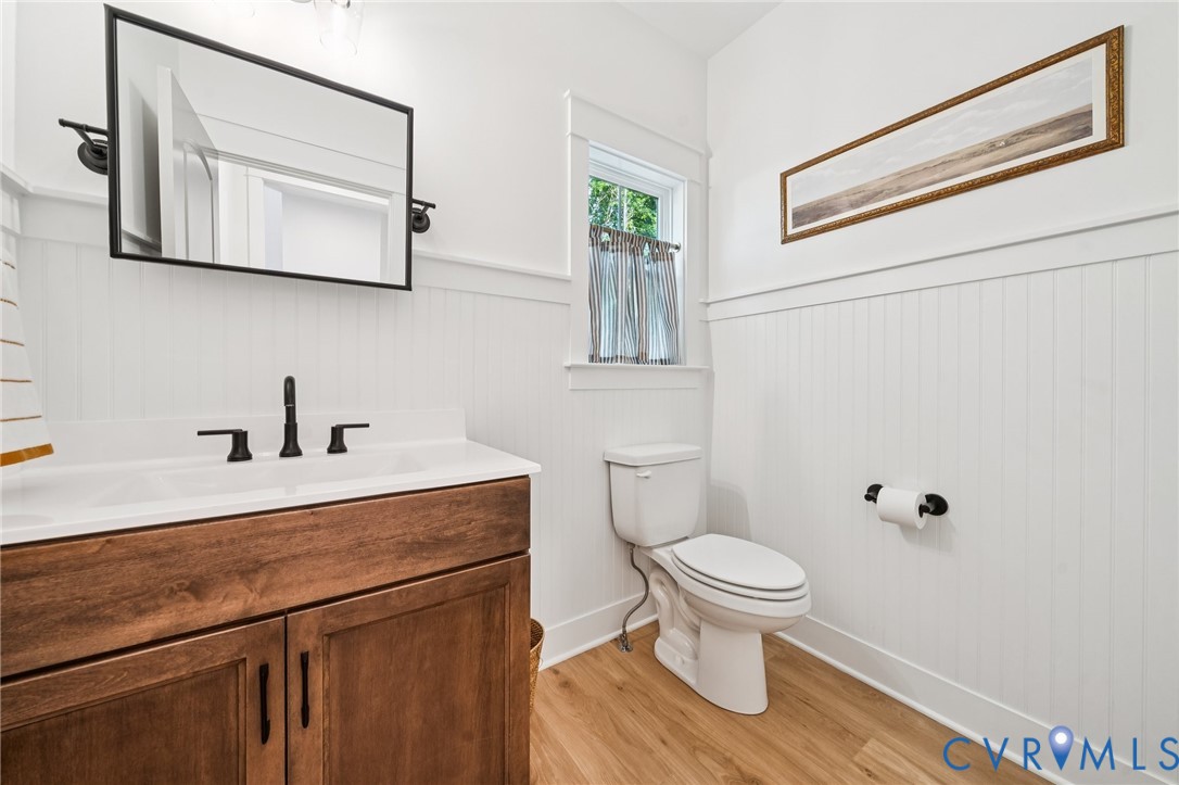 Bathroom featuring beadboard wall paneling, wood-finish flooring, a vanity with a white countertop and integrated sink, matte black hardware, and a framed mirror Bathroom featuring beadboard wall paneling, wood-finish flooring, a vanity with a white countertop and integrated sink, matte black hardware, and a framed mirror