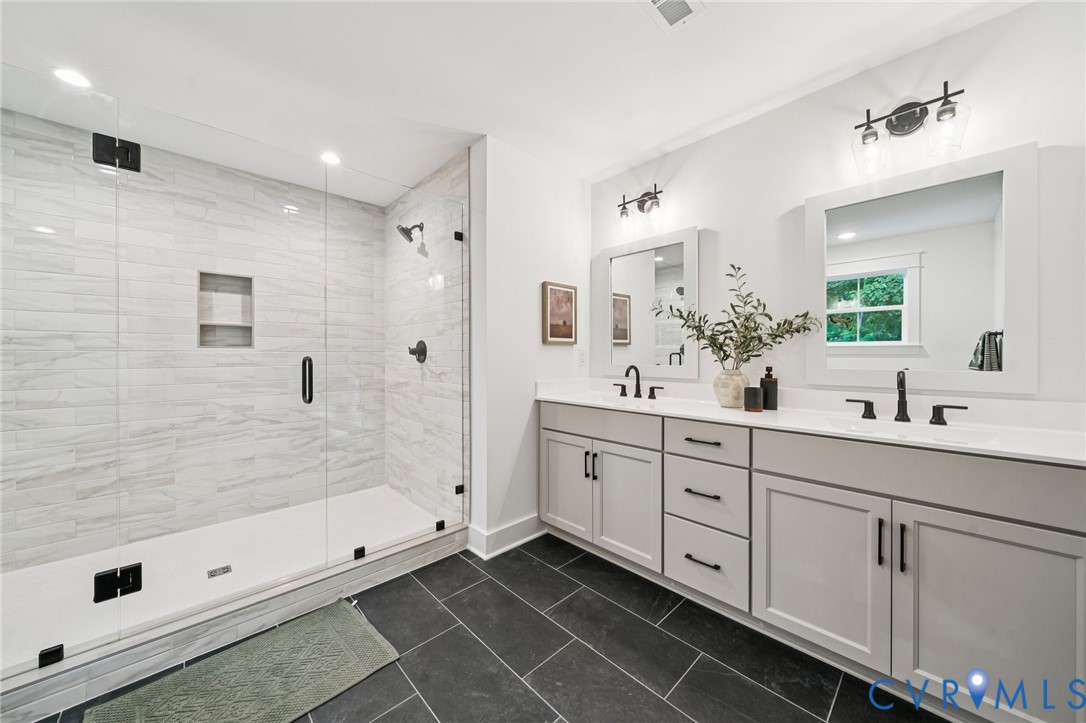 Double vanity with white countertops, light grey shaker-style cabinetry, matte black hardware, glass-enclosed walk-in shower with horizontal wall tile, and dark stone-finish floor tile