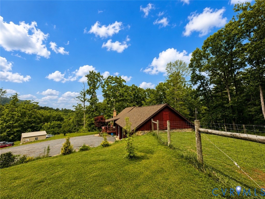 Rustic structure with red siding and a shingled roof, set on a sprawling lot with established landscaping, mature trees, a gravel driveway, and perimeter wire fencing