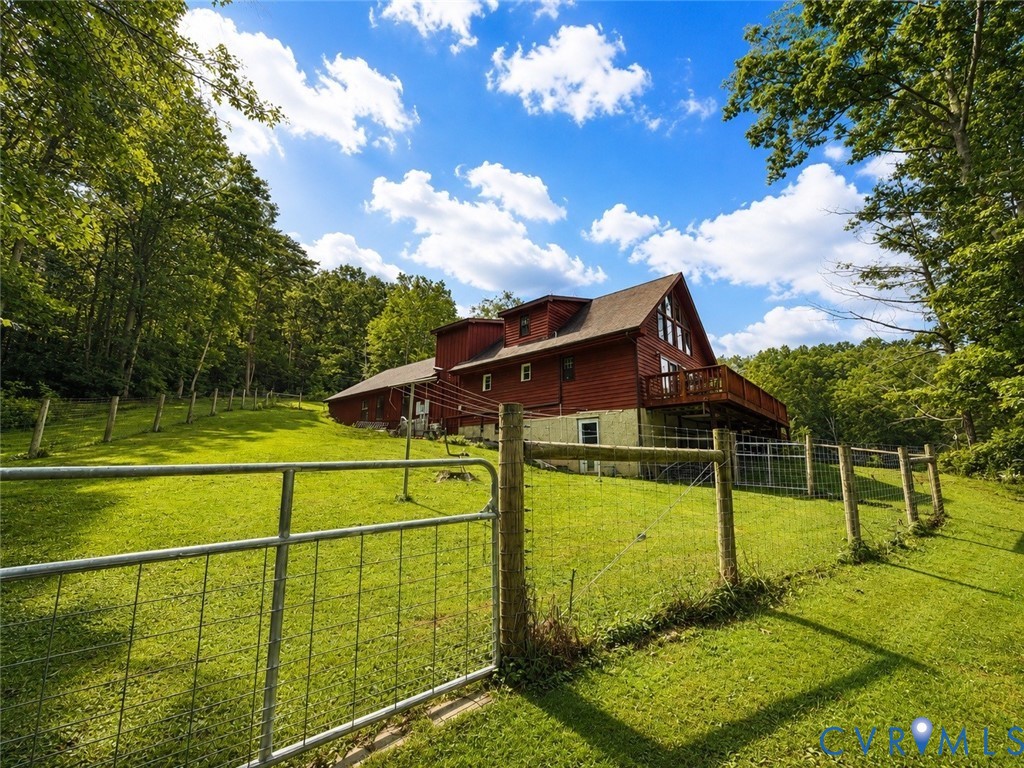 Multi-level residence with red siding and an elevated wood deck, situated on expansive grounds featuring fenced pasture areas and mature timber