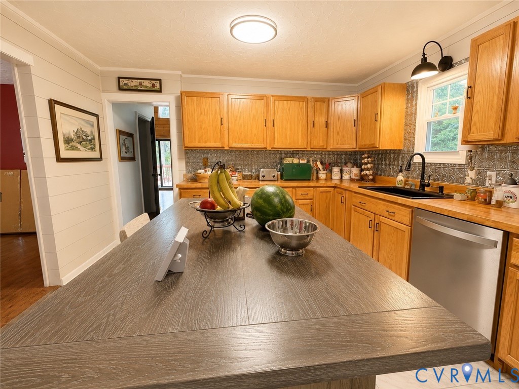 Kitchen featuring wood cabinetry, a central island with a wood-finish surface, a black drop-in sink, stainless steel dishwasher, and textured tile backsplash