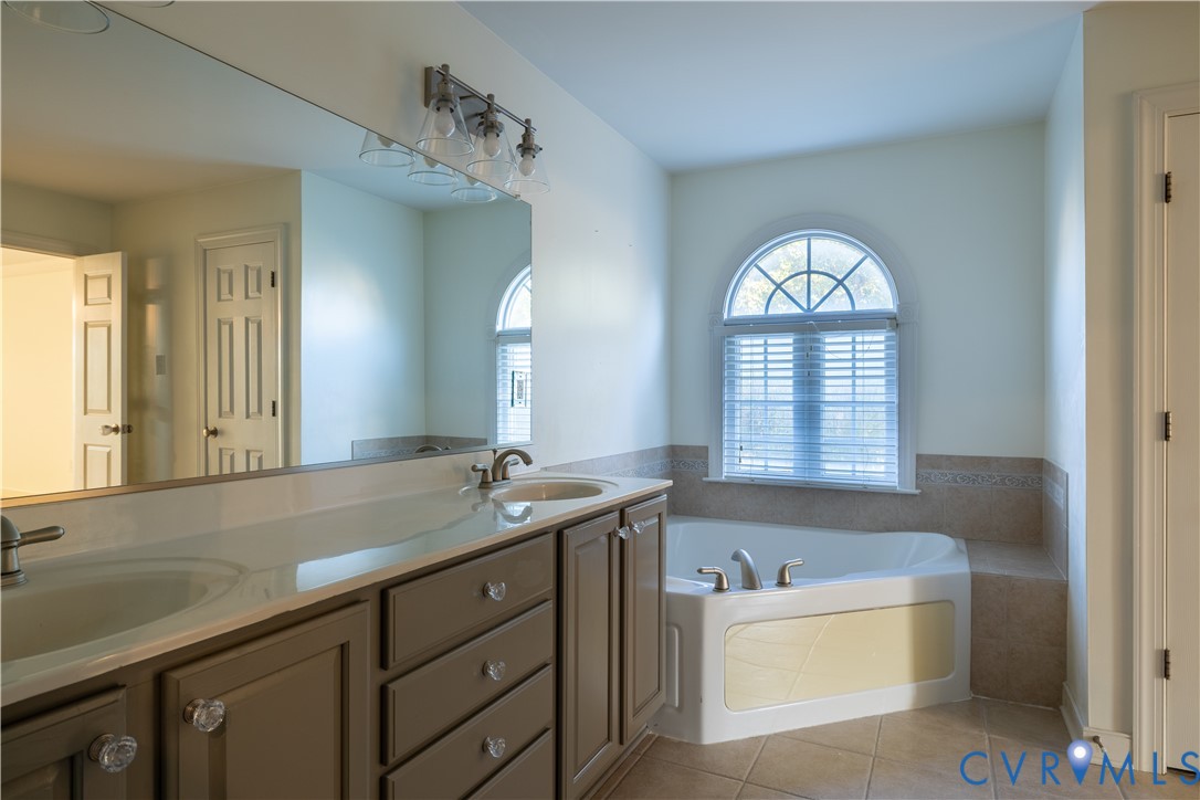 Dual-sink vanity with neutral cabinetry, expansive wall-to-wall mirror, corner soaking tub, arched window, and ceramic tile flooring