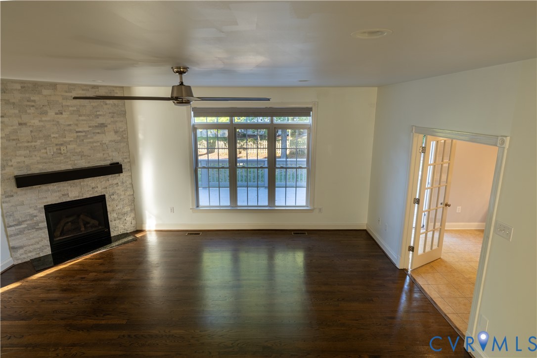 Living area featuring dark wood-finish flooring, a stacked stone fireplace surround with a linear mantel, multi-pane window configuration, and a ceiling fan