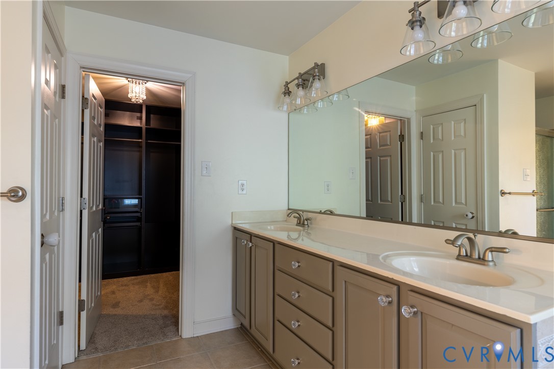 Dual-sink vanity with light-tone cabinetry, expansive wall-to-wall mirror, polished metal fixtures, and integrated storage, featuring a recessed walk-in closet with custom built-in shelving