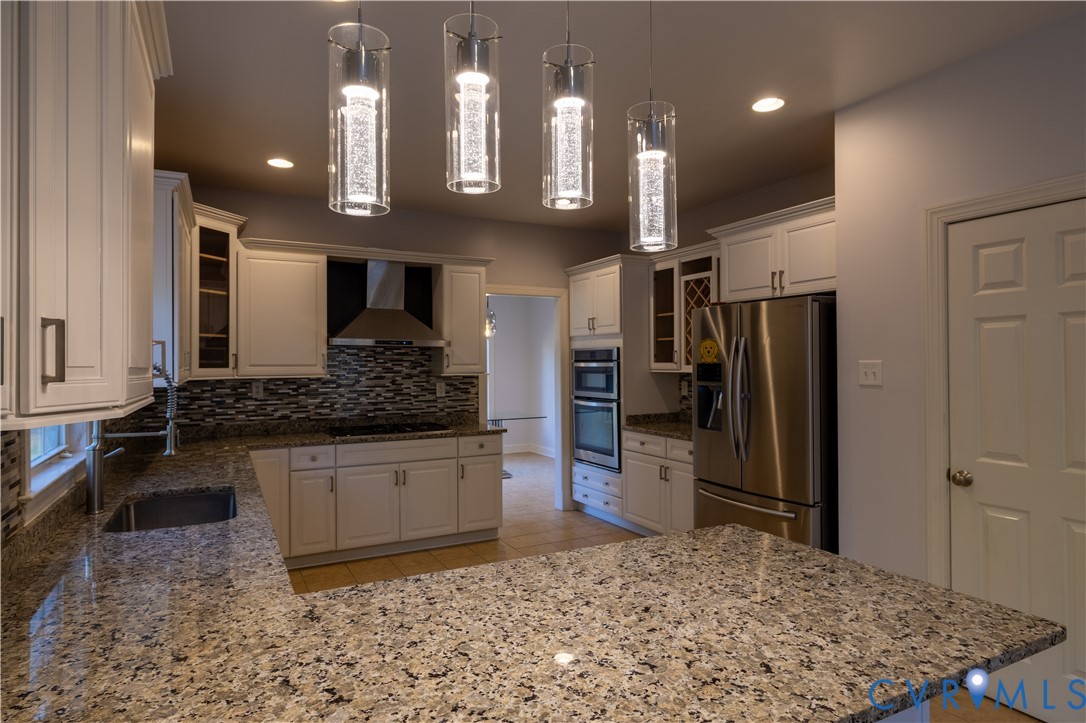 Kitchen featuring white cabinetry, stone countertops, stainless steel appliances, multi-textured stone tile backsplash, and four cylindrical pendant lights