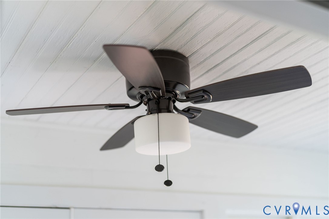 Matte black five-blade ceiling fan with integrated cylindrical frosted glass light fixture and bead-chain pull switches, mounted to a white beaded-plank ceiling