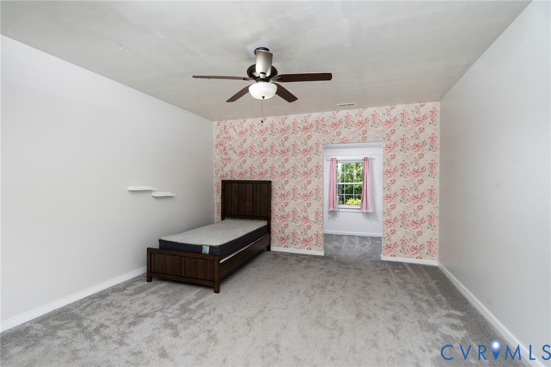 Room featuring carpeted flooring, decorative wallpaper accent wall, wall-mounted shelving, and a ceiling-mounted light fixture with a fan