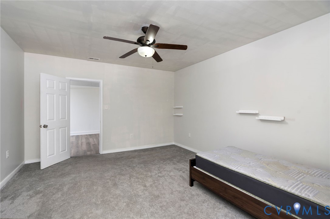 Neutral interior room featuring wall-to-wall carpeting, a multi-blade ceiling fan with integrated lighting, and white floating wall shelves