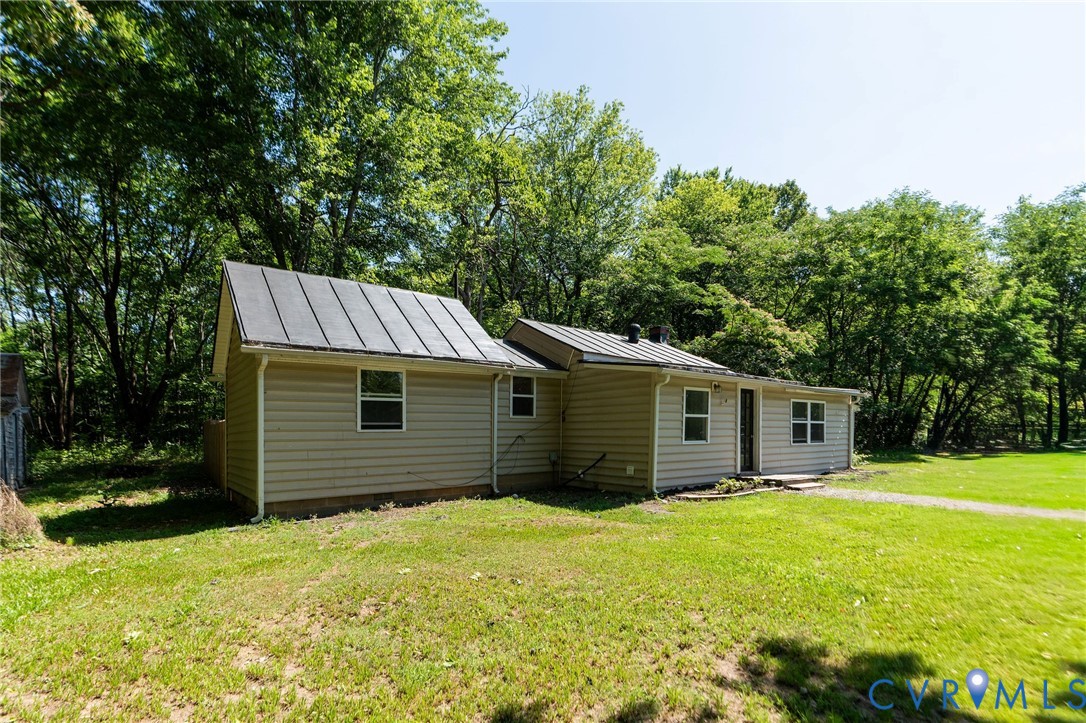 Single-story exterior featuring light-toned horizontal siding, a dark metal roof, and multiple window placements on a large grass lot with mature trees