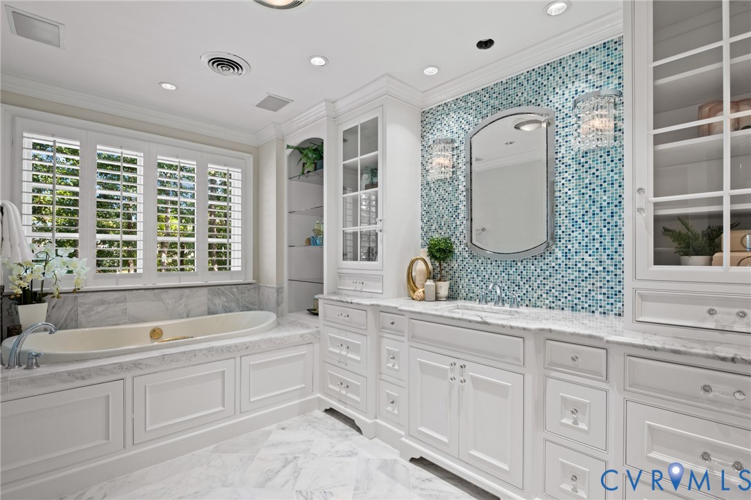 Custom cabinetry with glass-fronted displays, tiled mosaic accent wall, stone-surround soaking tub, stone tile flooring, and integrated recessed lighting