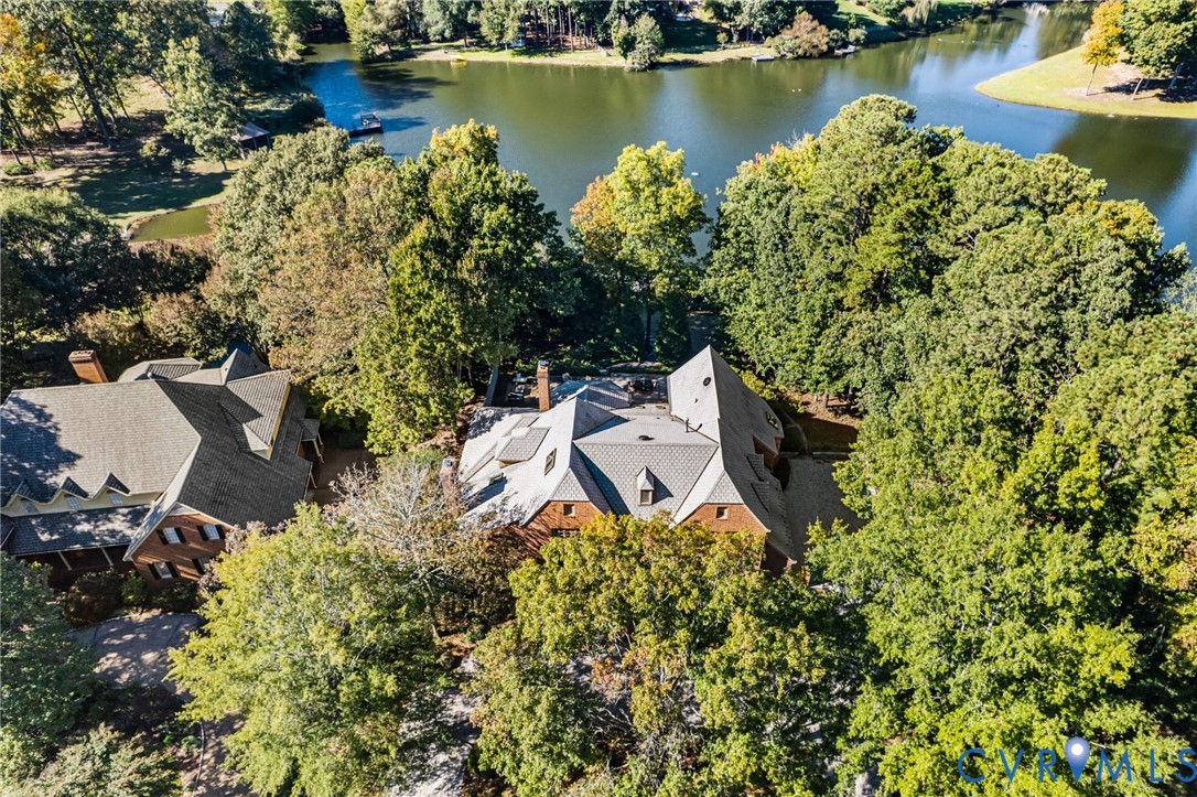 Aerial perspective of a multi-gabled brick residence featuring a multi-layered roofline, stone chimney, and expansive wooded grounds situated along a lakefront