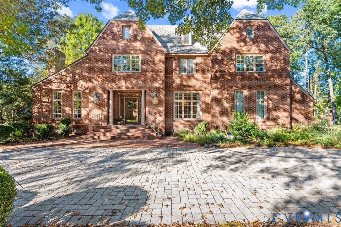 Two-story brick exterior featuring a steeply pitched roof, multi-pane windows, a columned entryway, and a paved stone driveway