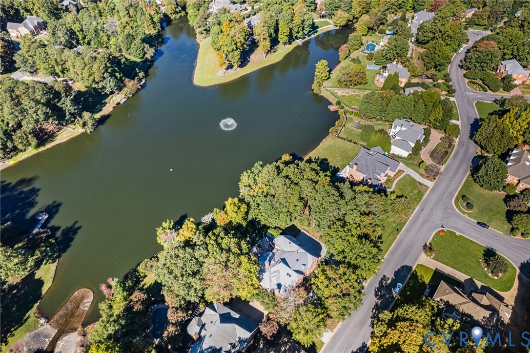 Aerial perspective of residential properties bordering a lake with a central fountain, featuring mature trees, landscaped lawns, and paved thoroughfares