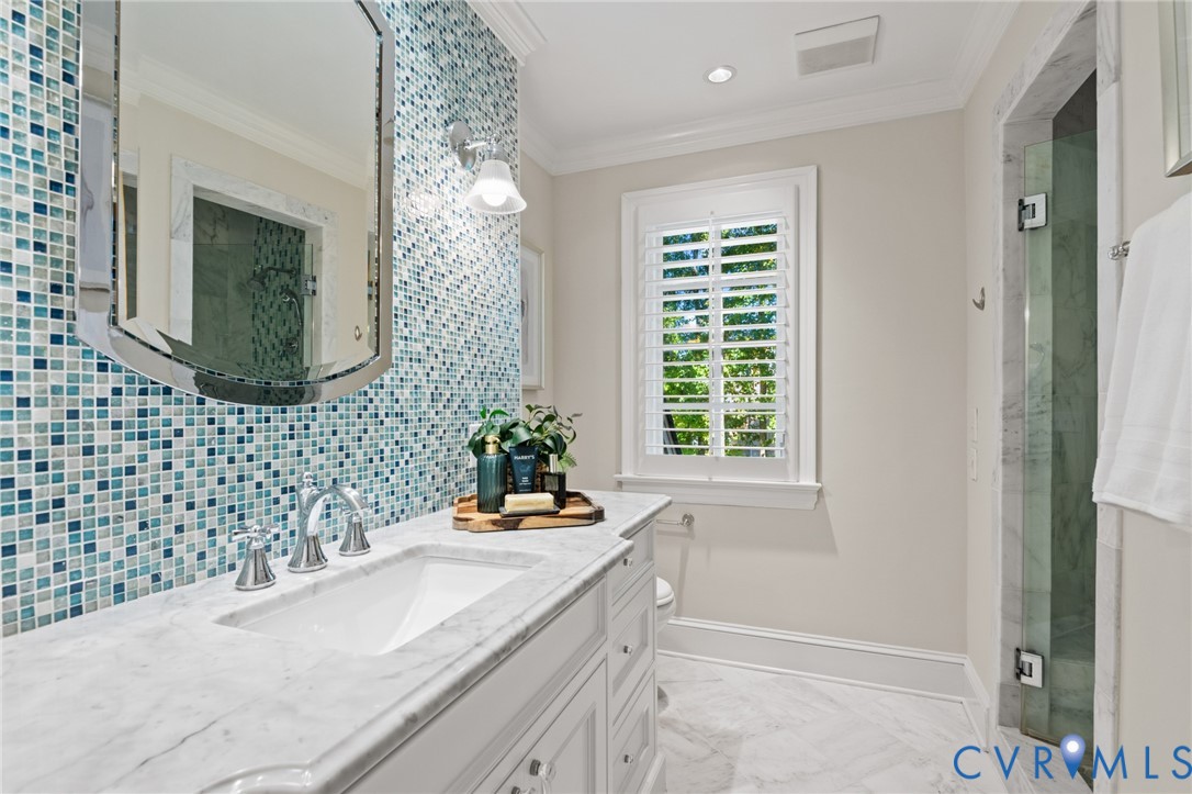 Custom vanity featuring a natural stone countertop, undermount sink, and polished chrome hardware, accented by a blue glass mosaic tile backsplash, framed wall mirror, and white plantation shutters