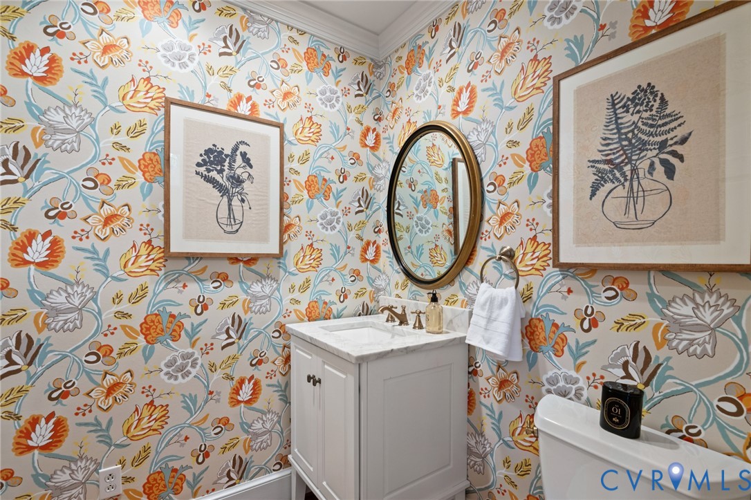 Powder room featuring floral patterned wallpaper, white vanity with stone countertop, oval gold-framed mirror, and metallic faucet hardware