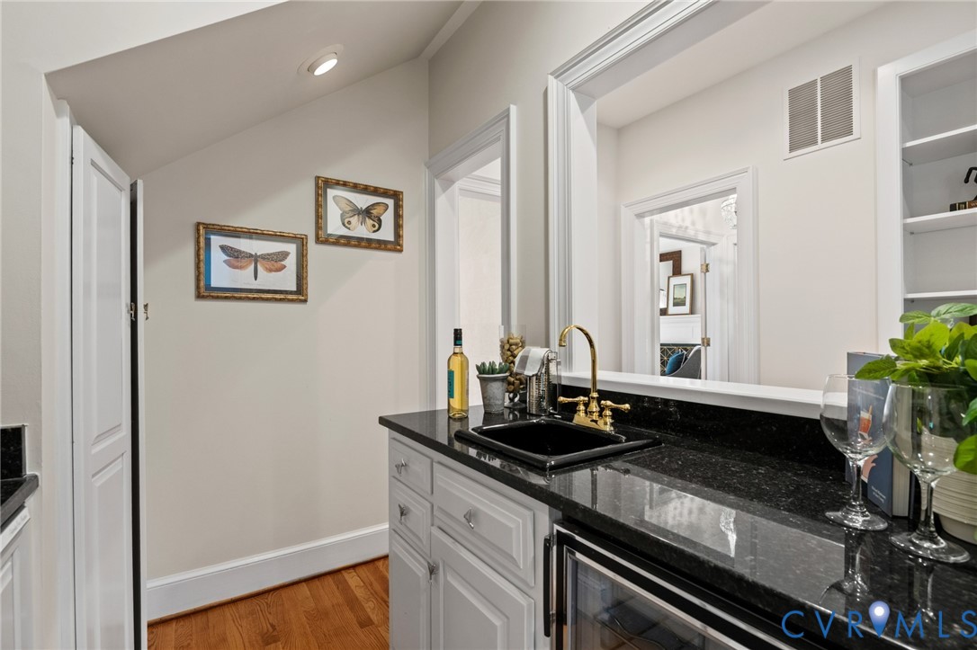 Wet bar featuring dark stone countertops, a black undermount sink with brass finish hardware, custom white cabinetry, and built-in shelving