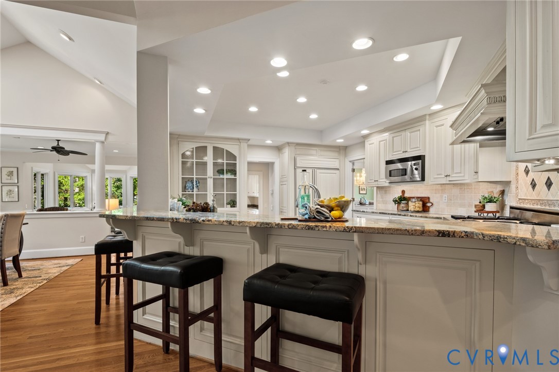 Kitchen featuring a multi-level stone countertop, custom white cabinetry, built-in glass-front display hutch, recessed lighting, and wood-finish flooring