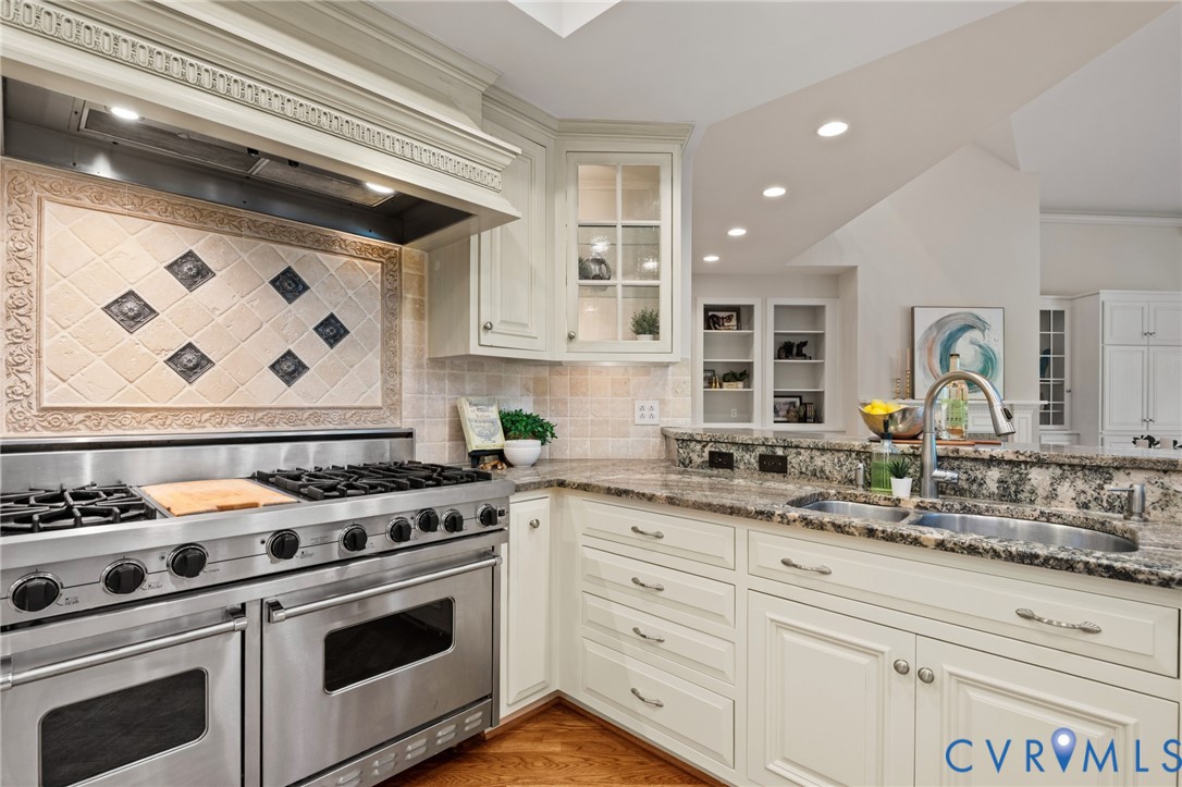 Kitchen featuring a stainless steel dual-oven gas range with a decorative tile backsplash, stone countertops, custom cabinetry with glass-front display sections, and a dual-basin undermount sink