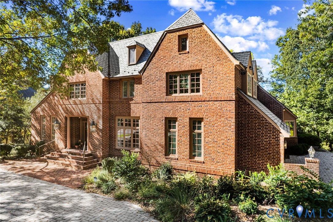 Tudor-style brick exterior featuring a steep-pitched roofline, multi-pane casement windows, stone-paved motor court, and detailed masonry facade