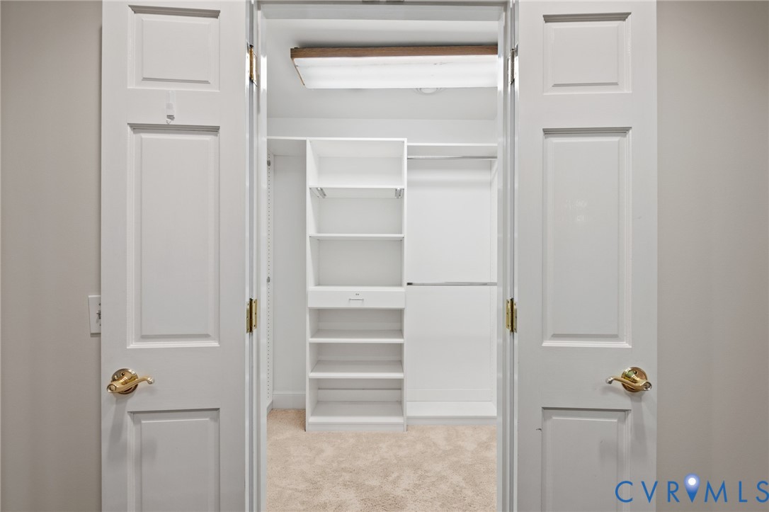 Walk-in closet featuring white built-in shelving, a dedicated drawer unit, dual hanging rods, and neutral carpeting, enclosed by white paneled double doors with brass-finish hardware