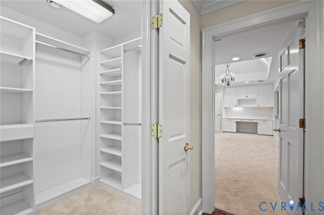 Walk-in closet featuring white custom built-in shelving, hanging rods, and carpeted flooring, opening to a space with built-in cabinetry and a desk area