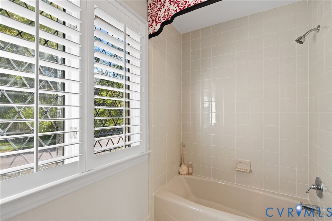 Bathtub with square tile surround, polished chrome fixtures, and window featuring white plantation shutters and leaded glass accents