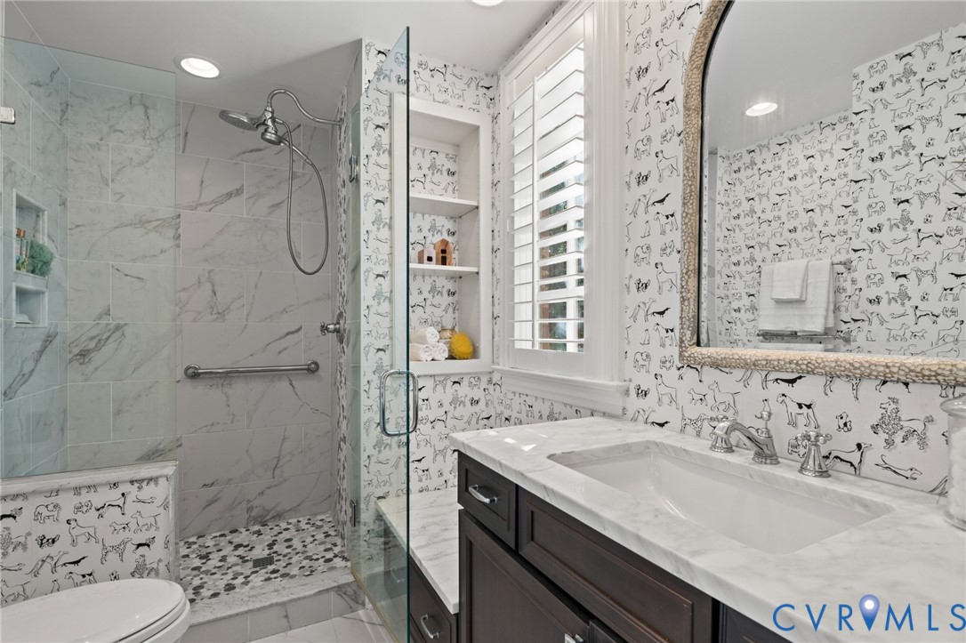 Bathroom featuring stone-finish wall tile, a walk-in shower with a pebble-style floor, a built-in storage niche, stone-top vanity, and plantation shutters