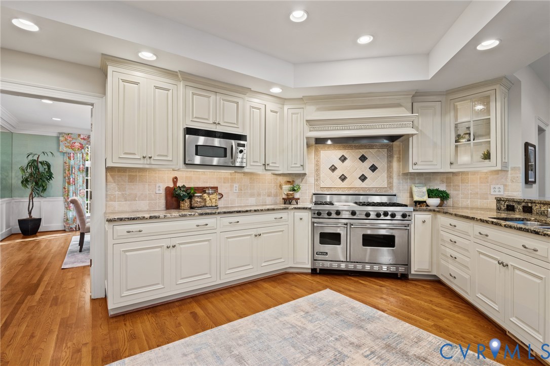 Kitchen featuring off-white cabinetry, stone-finish countertops, tile backsplash with decorative inset, stainless steel double oven range, and wood-finish flooring