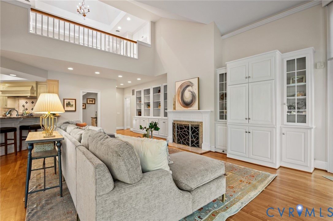 Vaulted ceilings with mezzanine overlook, fireplace with carved mantle, extensive built-in cabinetry with glass display panels, recessed lighting, and wood-finish flooring