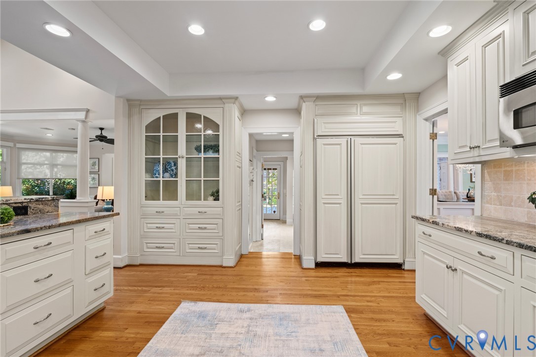 Custom off-white cabinetry featuring glass-paneled display doors, integrated panel-front refrigerator, stone countertops, recessed ceiling lighting, and wood-finish flooring