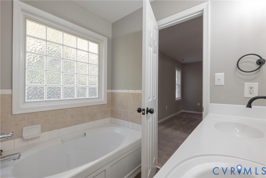 Soaking tub with tile surround, glass block privacy window, dual-basin vanity with solid-surface countertop, and matte black hardware fixtures