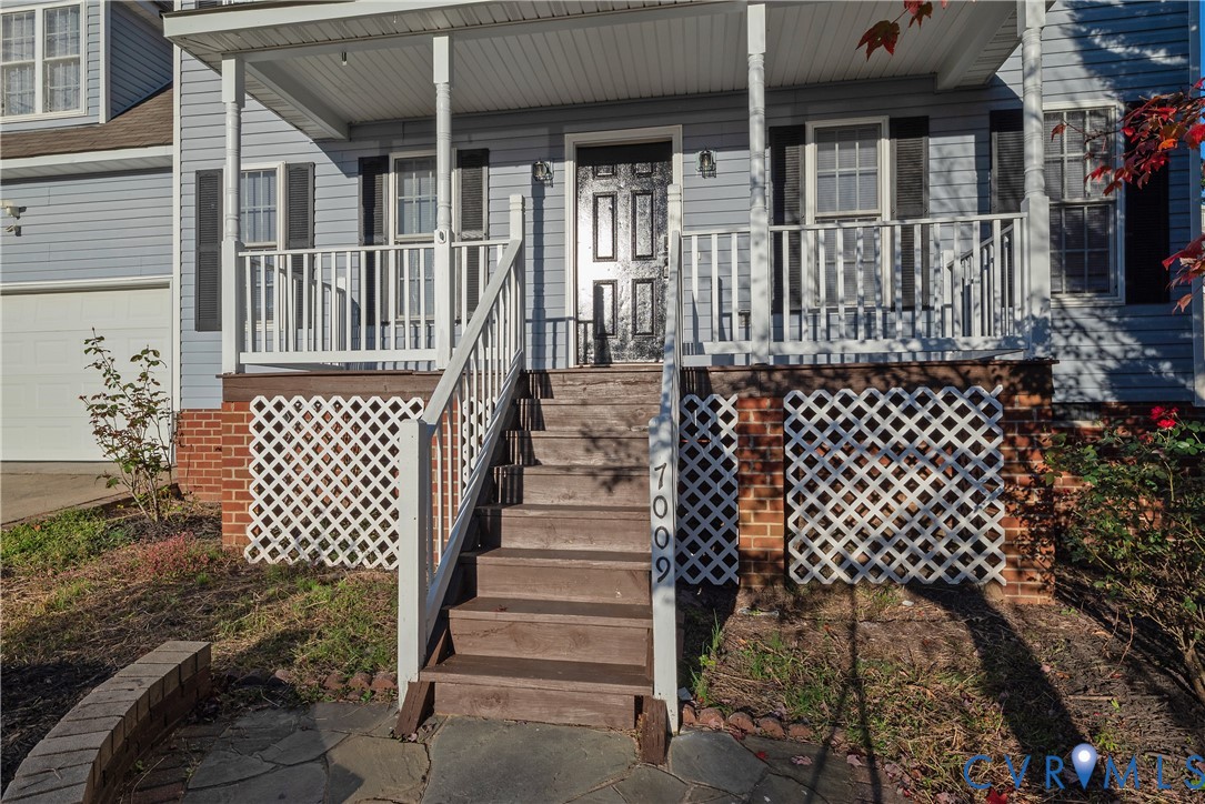 Front exterior featuring blue horizontal siding, a covered porch with white railing, brick foundation skirting, wood-finish stairs, and a flagstone patio