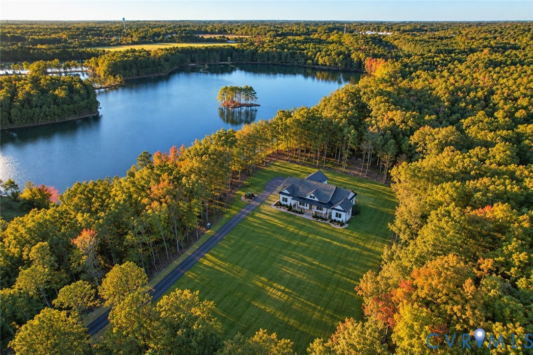 Aerial view of residential property featuring a sprawling lawn, a paved driveway, and substantial wooded acreage bordering a large pond Aerial view of residential property featuring a sprawling lawn, a paved driveway, and substantial wooded acreage bordering a large pond