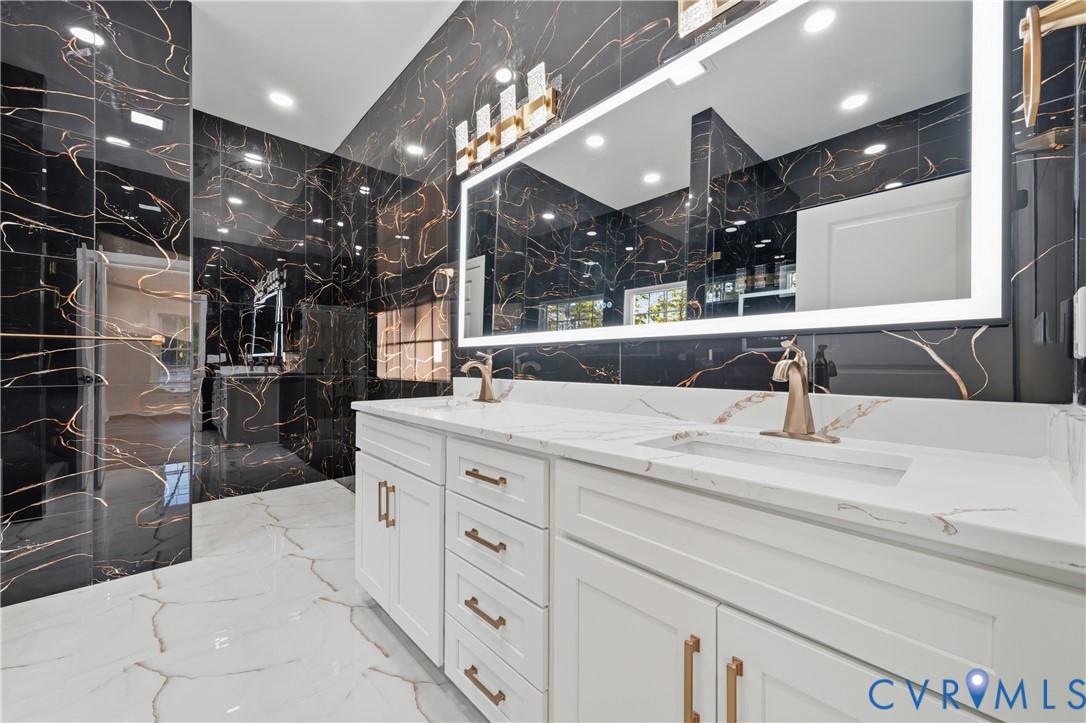 Bathroom featuring large-format dark veined wall tile, white cabinetry, quartz countertop with dual integrated sinks, metallic finish fixtures, and a rectangular LED-backlit mirror