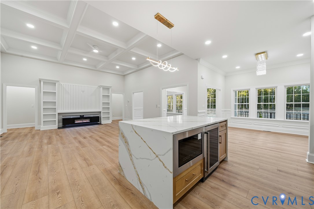 Coffered ceilings, center island with stone waterfall edges, built-in beverage refrigerator, wood-finish flooring, and integrated wall shelving with a built-in fireplace