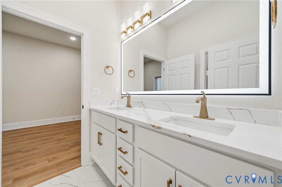 Dual-sink vanity with stone-look countertop, integrated rectangular basins, brushed gold-finish fixtures, oversized LED-backlit mirror, and white shaker-style cabinetry with matching gold-finish hardware