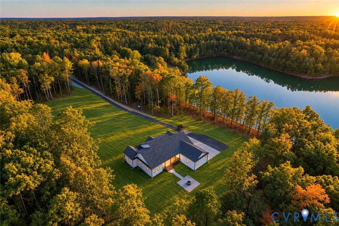 Aerial perspective of a sprawling property featuring extensive wooded acreage, a private shoreline along a body of water, a paved driveway, and a stone-paver patio with a built-in fire pit