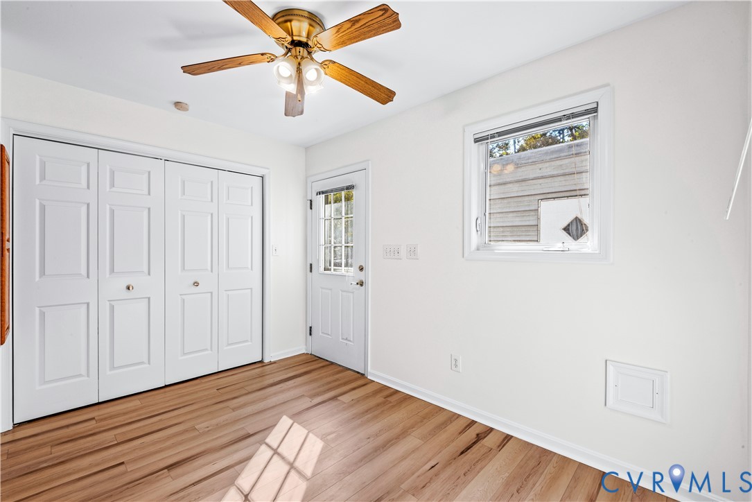 Wood-finish flooring, white four-panel bi-fold closet doors, brass-finish ceiling fan with integrated lighting, single-pane window, and white exterior entry door with glass pane