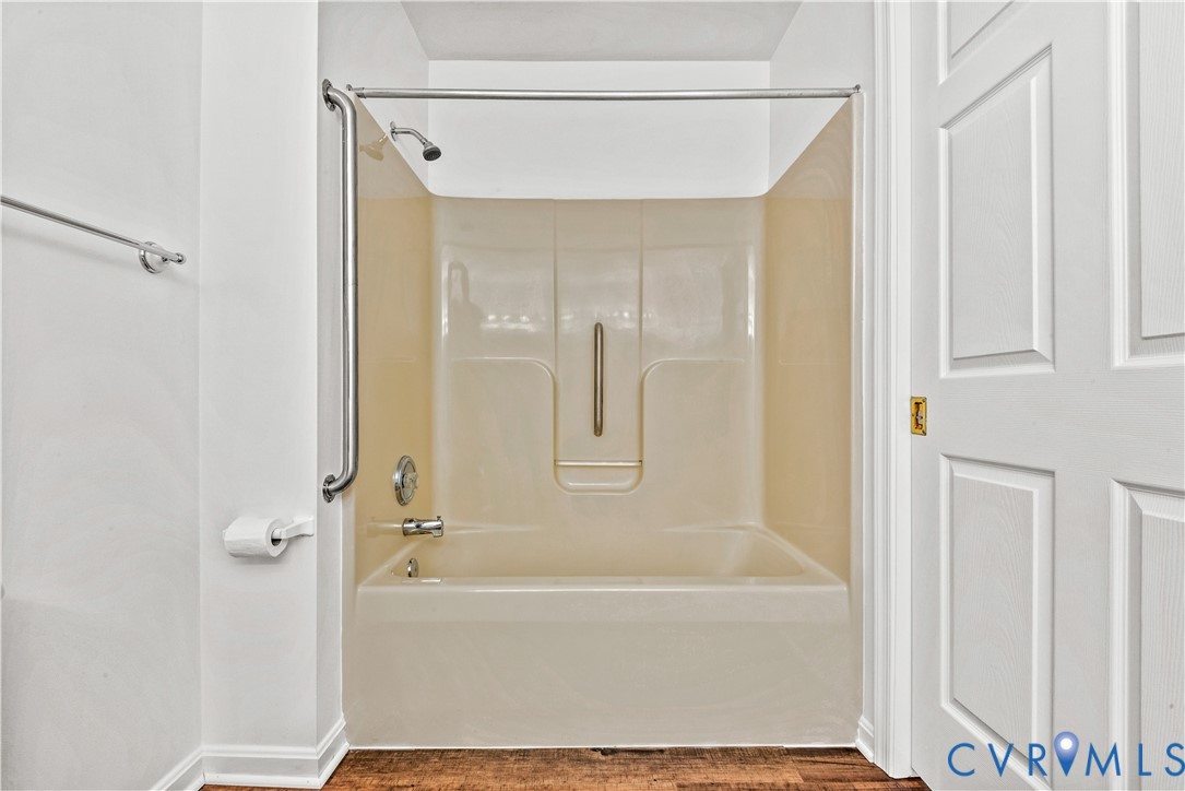 Tub and shower combination with an integrated surround, metallic wall-mounted grab bars, wood-finish flooring, and a multi-panel door with gold-tone hardware