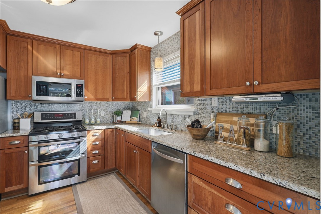 Kitchen featuring wood cabinetry, stone countertops, mosaic tile backsplash, stainless steel appliances, and a pendant light fixture
