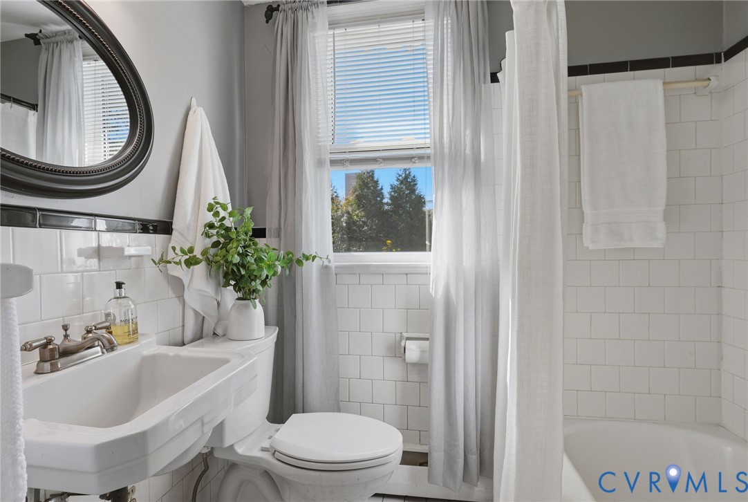 Wall-mounted porcelain sink with metallic dual-handle faucet, oval-framed mirror, white subway tile wall surround, and deep soaking tub