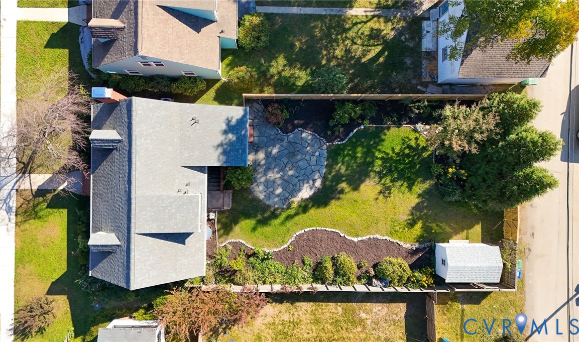 Aerial view of a rear yard featuring a stone patio, manicured lawn, perimeter landscaping beds, and a detached storage structure enclosed by fencing
