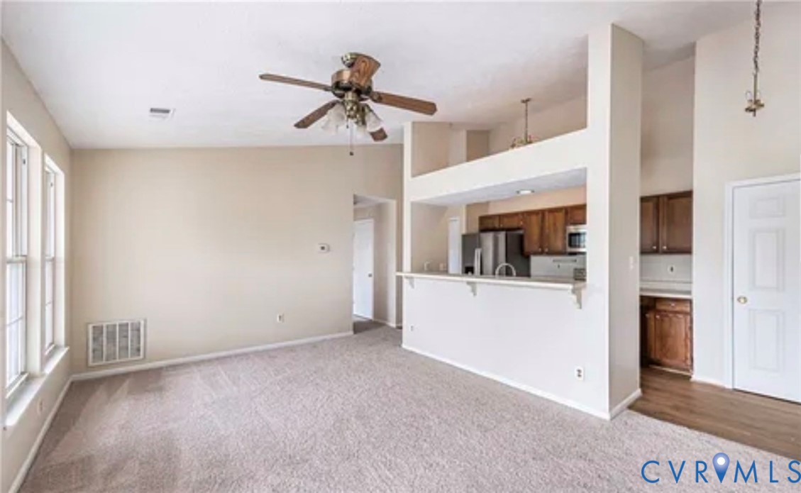 Vaulted ceilings featuring a ceiling fan, pass-through kitchen breakfast bar, wood-finish cabinetry, and neutral carpeting