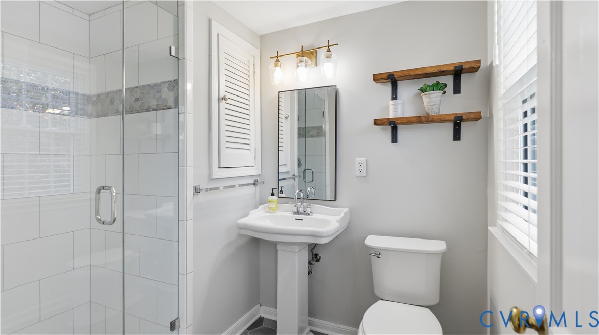 Bathroom featuring a glass-enclosed shower with subway tile surround, white pedestal sink, three-light vanity fixture, and wall-mounted wooden shelving