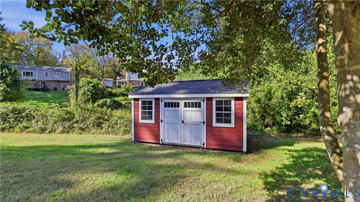 Exterior storage structure with a red wood-sided facade, asphalt shingle roof, double-entry doors, and window accents situated on a grass-covered lot
