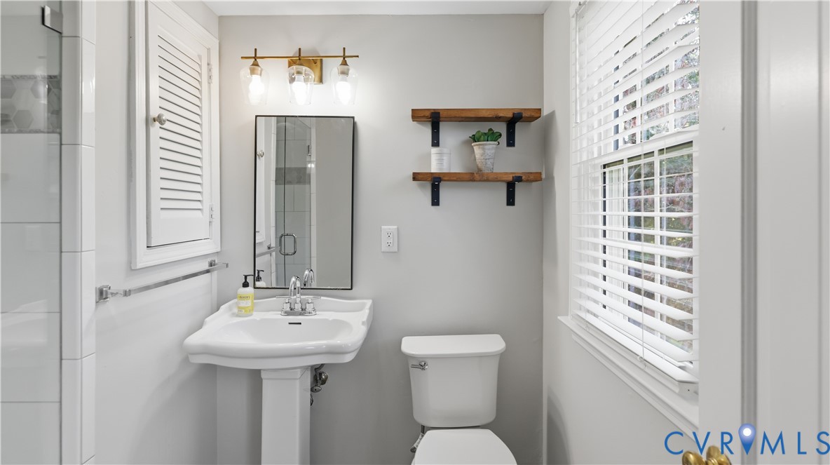 Bathroom featuring a pedestal sink, wall-mounted three-light fixture, framed rectangular mirror, built-in louvered-door cabinet, and wall-mounted wood shelving