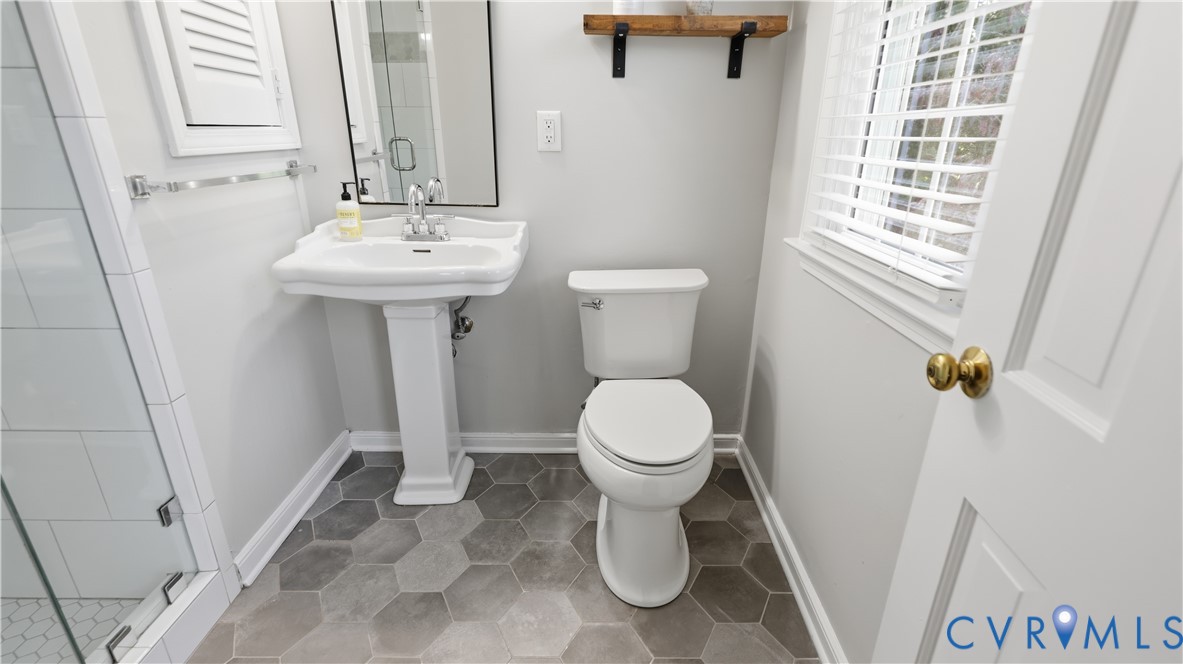 Bathroom featuring a white pedestal sink, glass-enclosed shower with white subway tiling, grey hexagonal floor tile, and a wall-mounted wooden shelf
