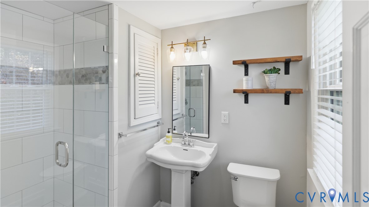 Bathroom featuring a walk-in shower with glass enclosure, white subway tile surround, pedestal sink, wall-mounted shutter-style cabinetry, and a three-light vanity fixture