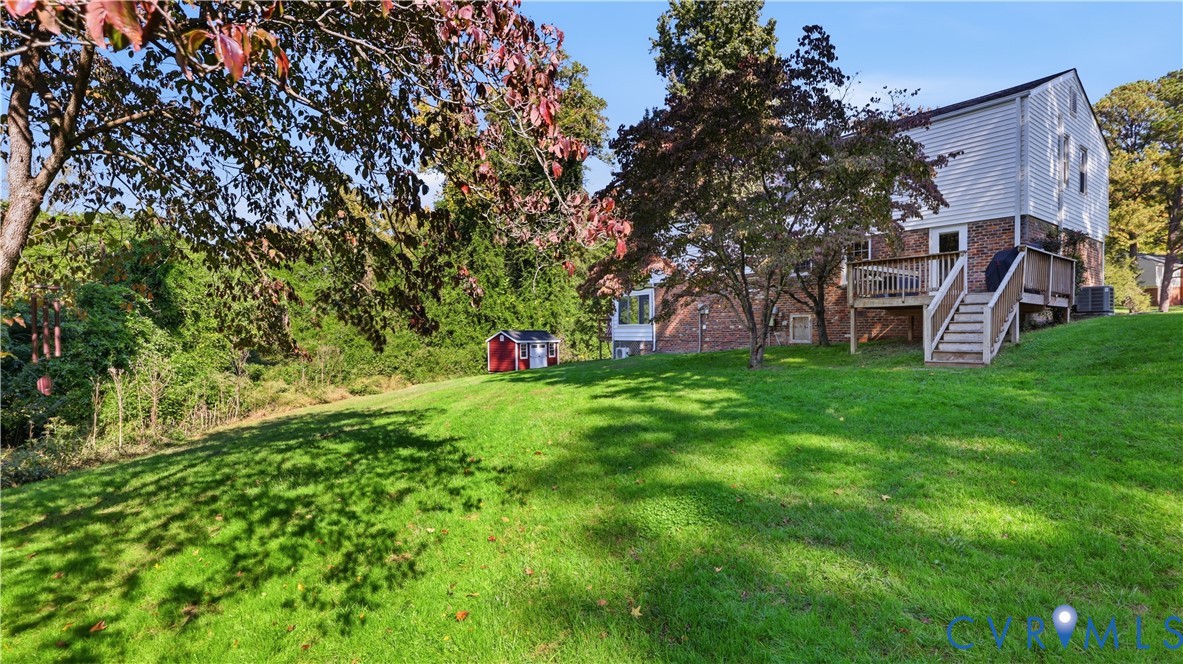 Exterior featuring a brick and siding facade, raised wood deck with staircase, expansive sloped lawn, mature trees, and detached storage shed